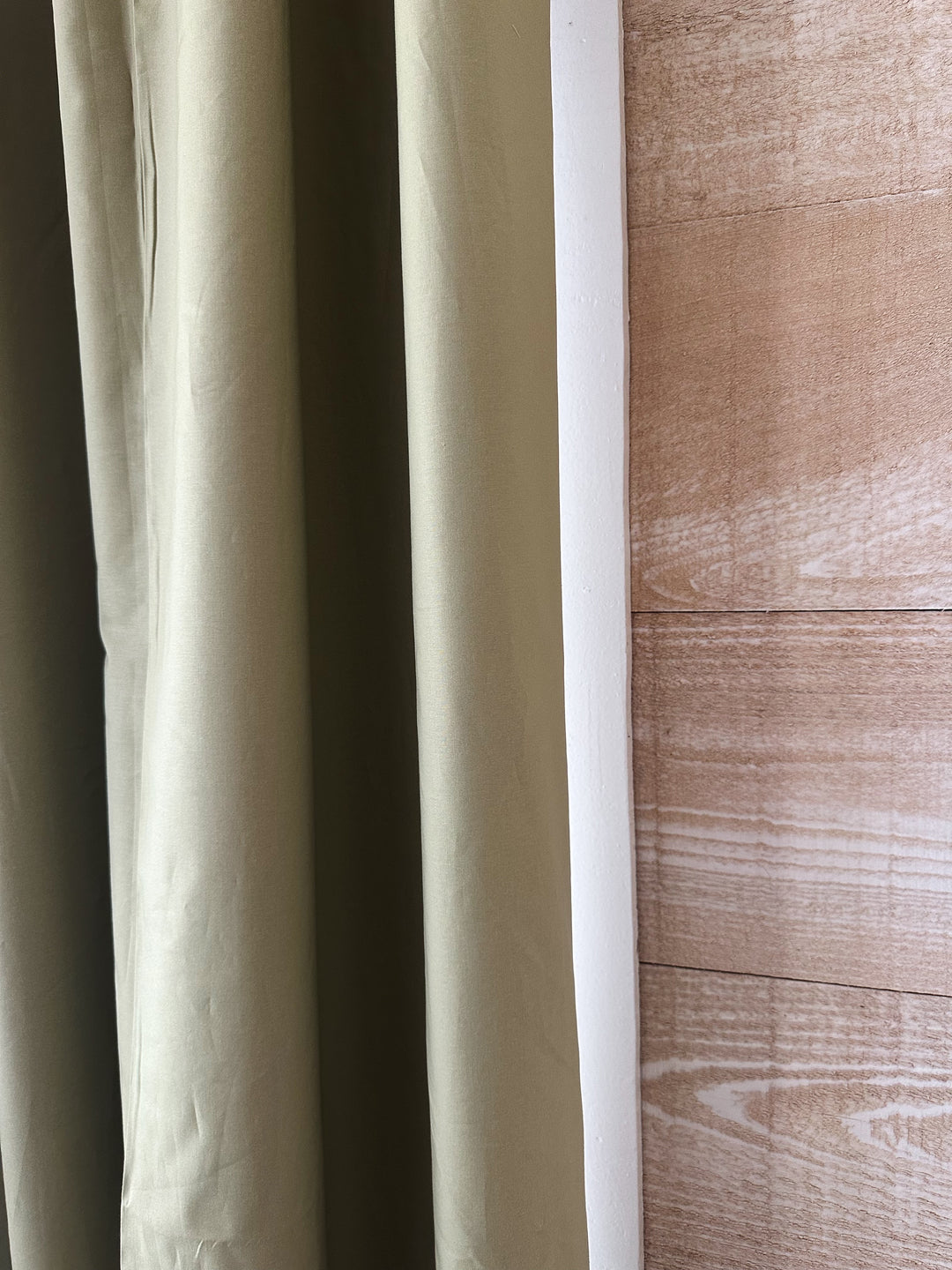 Edge of a sage green cotton panel hanging beside white window trim and a warm plank wall