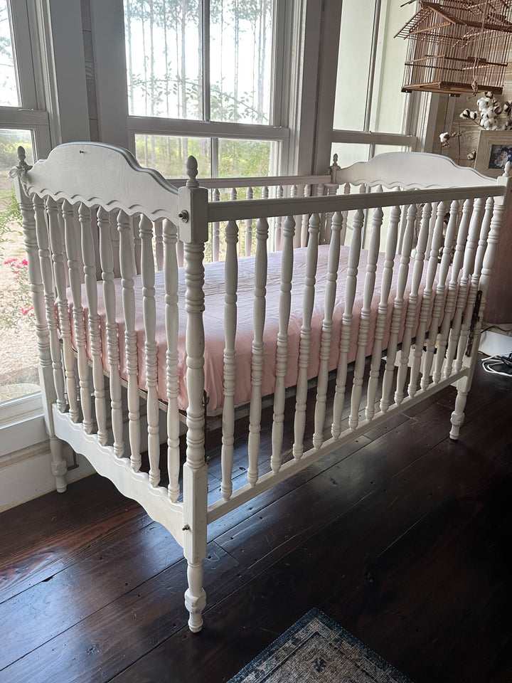 White spindle crib made up with a Sisters Pink fitted crib sheet beside tall windows, cotton stems and a hanging birdcage