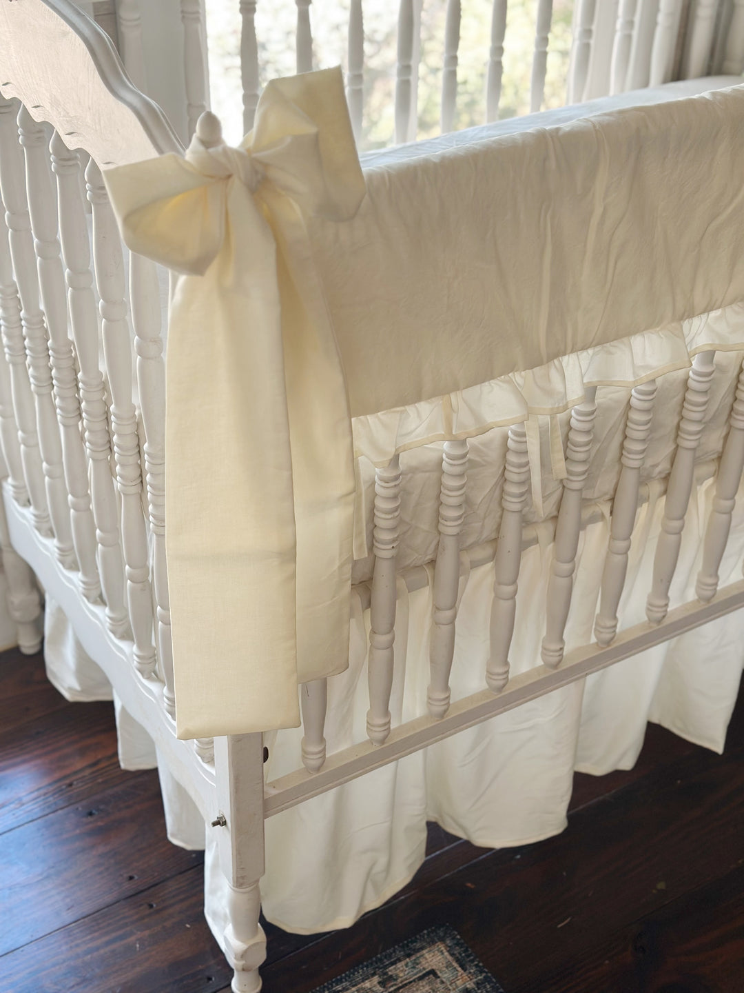 Close-up of an ivory cotton bow knotted at the crib's corner finial beside the ruffled ivory rail cover, gathered skirt below