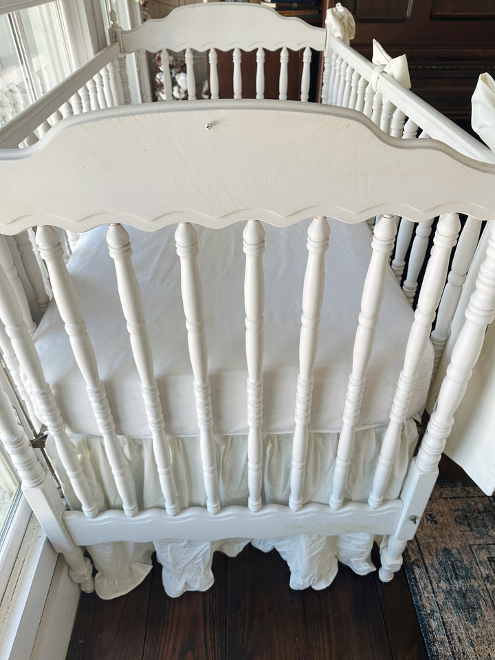 Head end of the white spindle crib with scalloped headboard, porcelain ruffled skirt gathered below the mattress