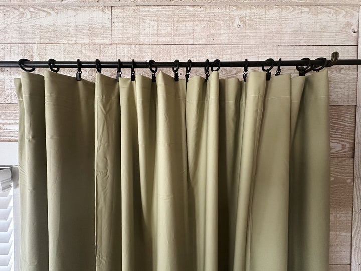 Header of the sage green curtains hung from black clip rings on an iron rod, soft folds falling from each clip