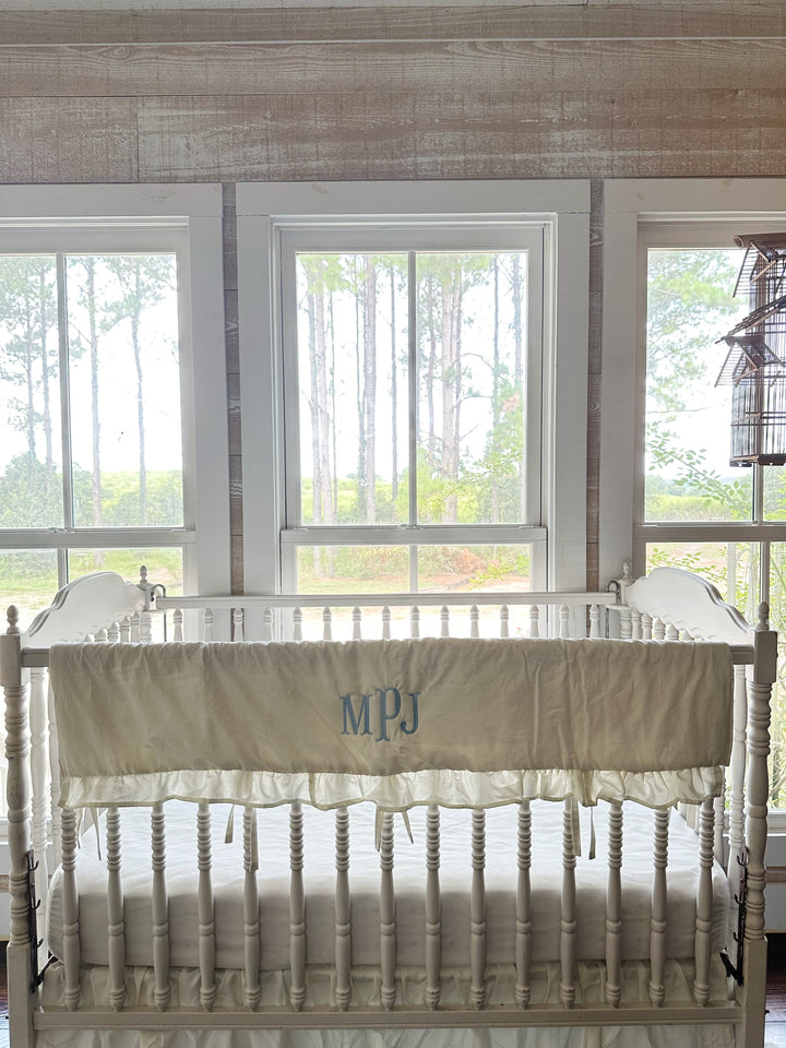 Porcelain ruffled crib rail cover with a blue monogram tied along the front rail of a white spindle crib