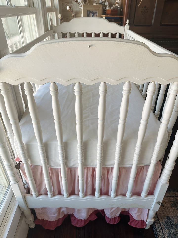 Head end of the white spindle crib with scalloped headboard, Sisters Pink and blush ruffled skirt below