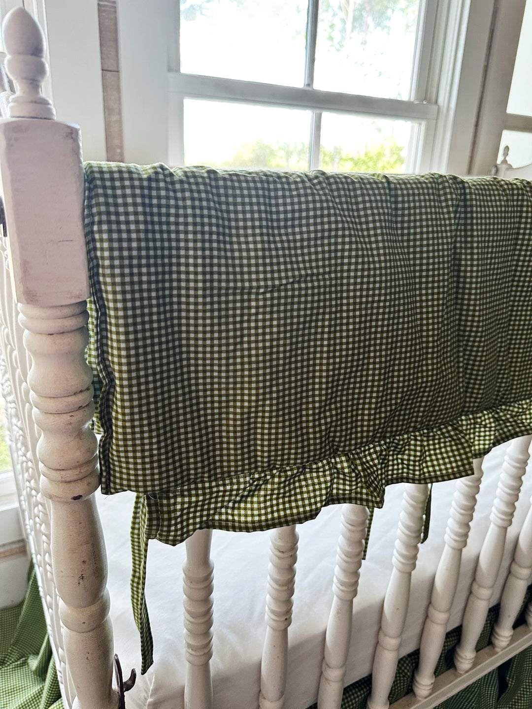 Green gingham crib rail cover tied at the corner post of a white crib beside a sunny window