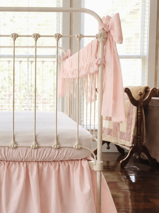 Looking down the crib rail at the pink bow tail and ruffle, with a patchwork quilt on a wooden rack behind