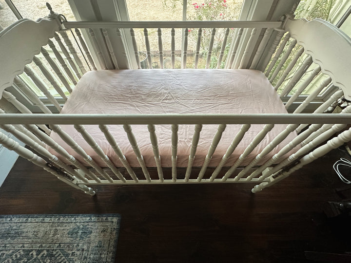 Overhead angled view of the Sisters Pink fitted crib sheet on a white turned-spindle crib above dark wood floors