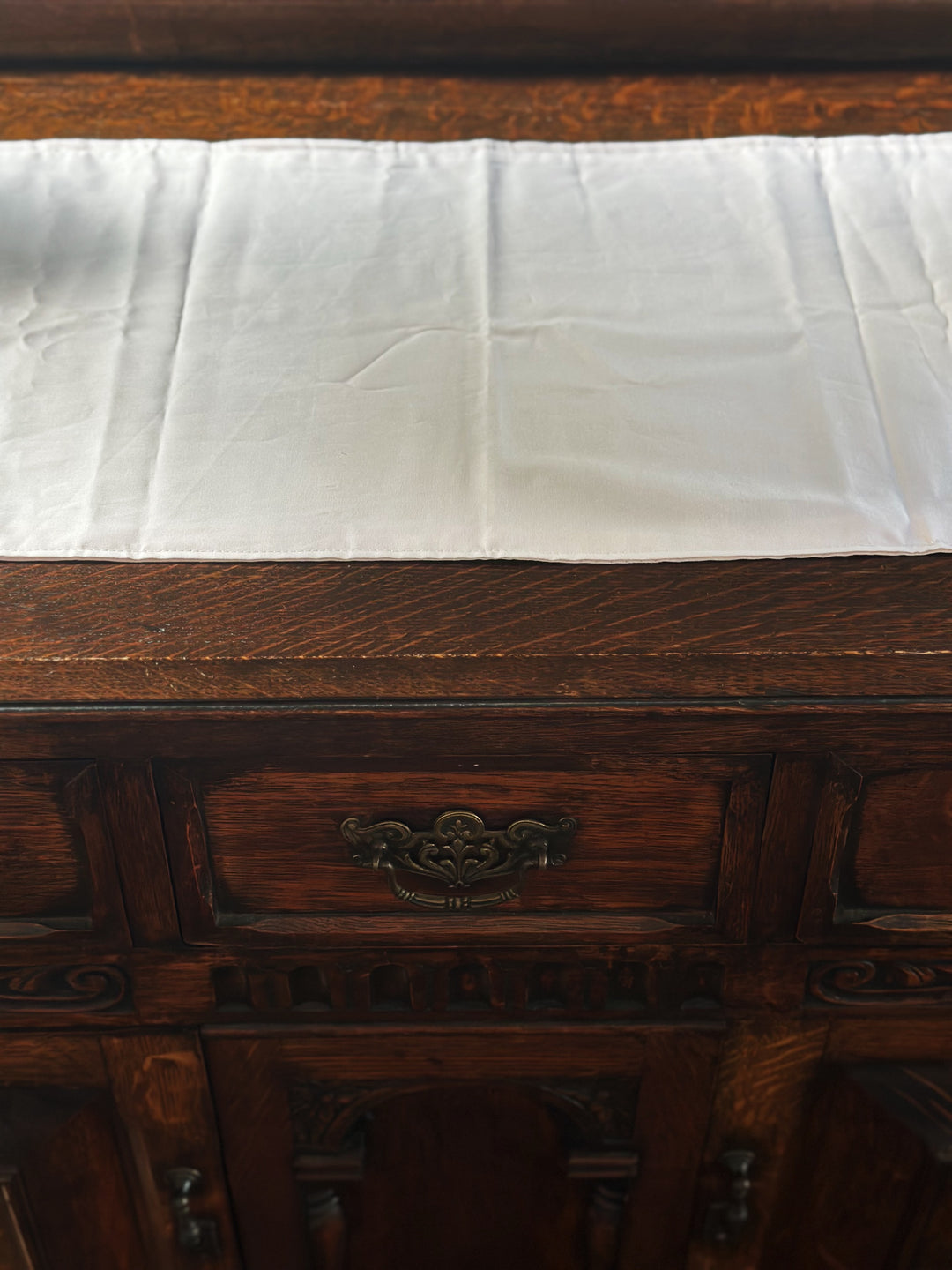 Baby pink runner across the top of a quarter-sawn oak dresser, above a drawer with an ornate brass pull