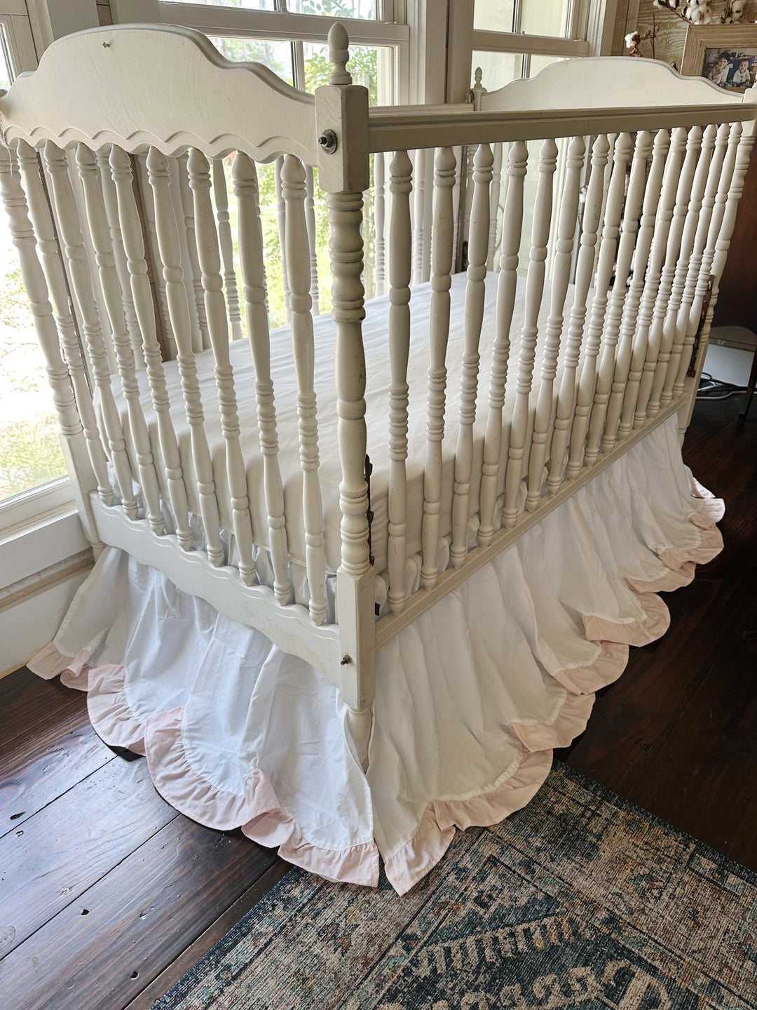 White scalloped crib skirt with a baby pink ruffle on a white spindle crib beside a sunny window