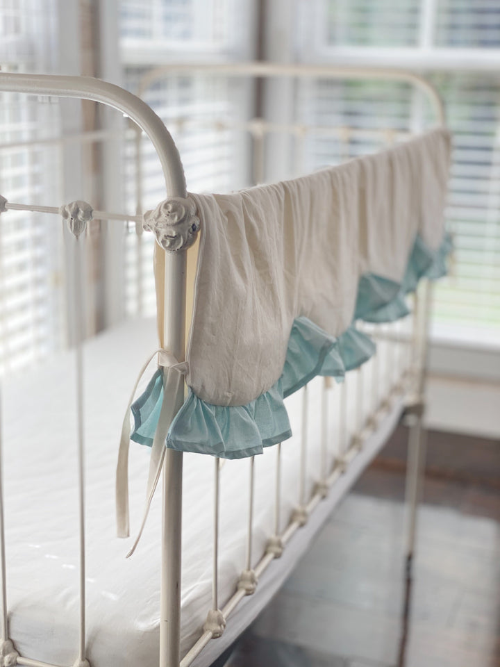 View down the length of an ivory crib rail cover with repeating scallops edged in mist blue