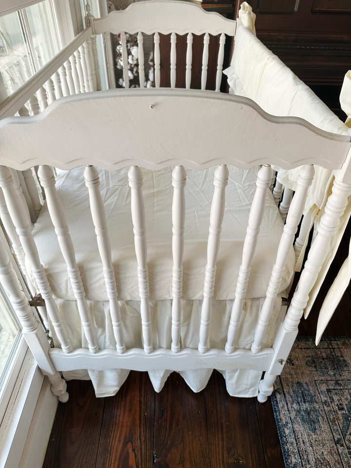 Close overhead view of the scalloped crib headboard with ivory skirt gathered below the mattress and the rail cover at the edge