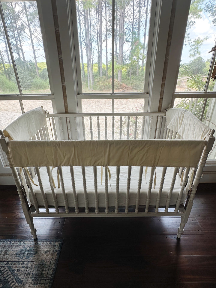 Long ivory crib rail cover tied along the front rail above a dark wood floor and patterned rug