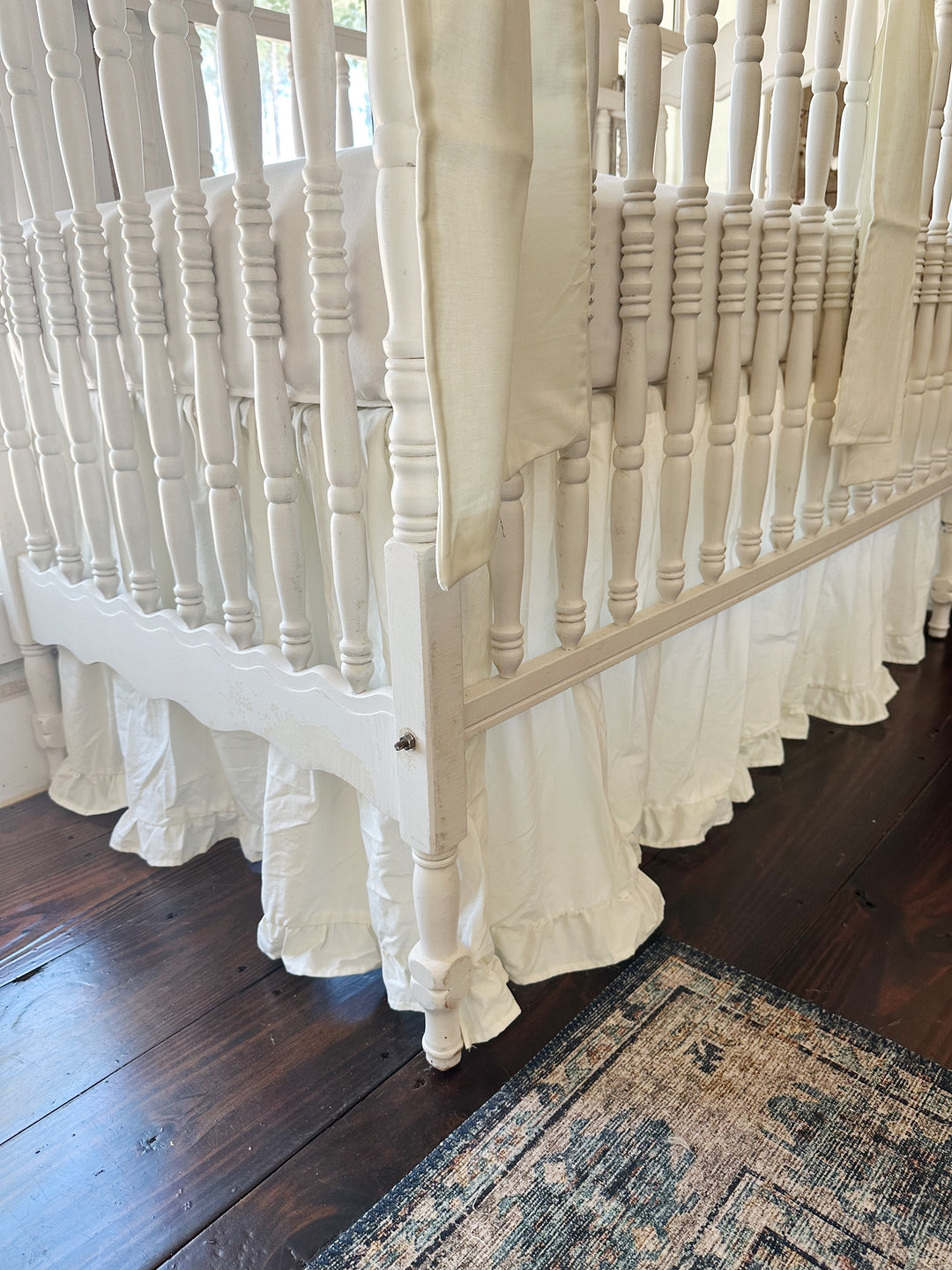 Crib corner with scalloped end rail and vintage rug, porcelain ruffled skirt falling in soft gathers to the floor