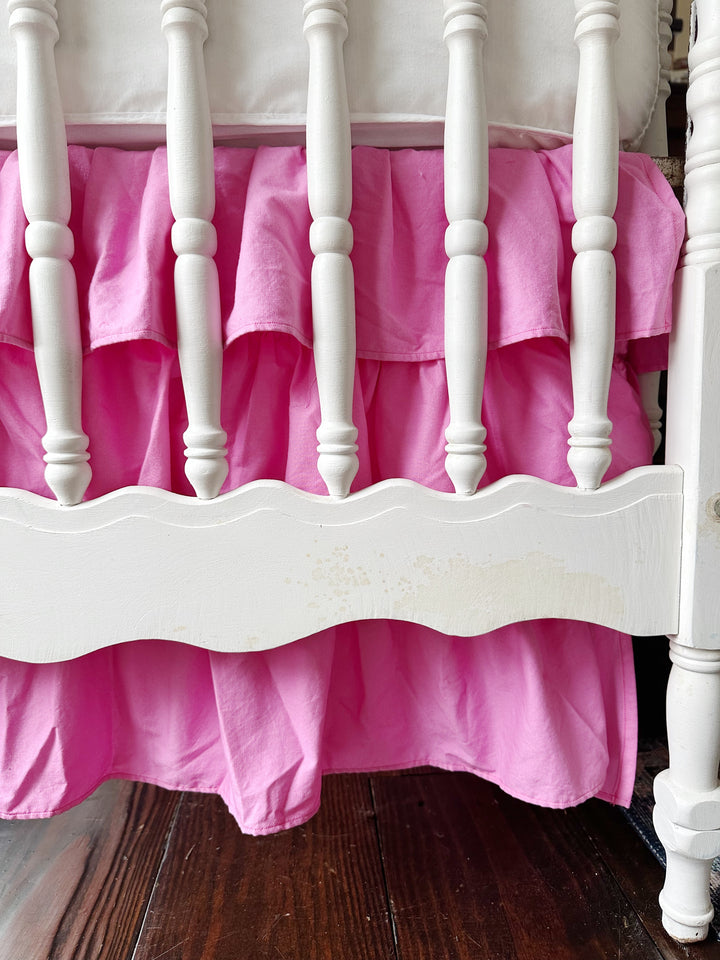 Close-up of the tiered pink ruffles at the end of a white crib with a scalloped rail, the hem falling just above the wood floor