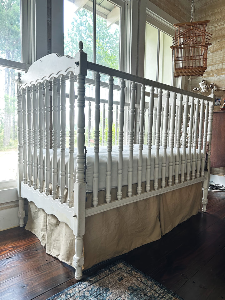 Side view of oatmeal tailored crib skirt on antique spindle crib with rustic wood walls - High Cotton Textile