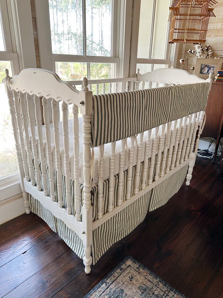 Three-quarter view of the crib, the ticking rail cover running the length of the far rail above the striped skirt
