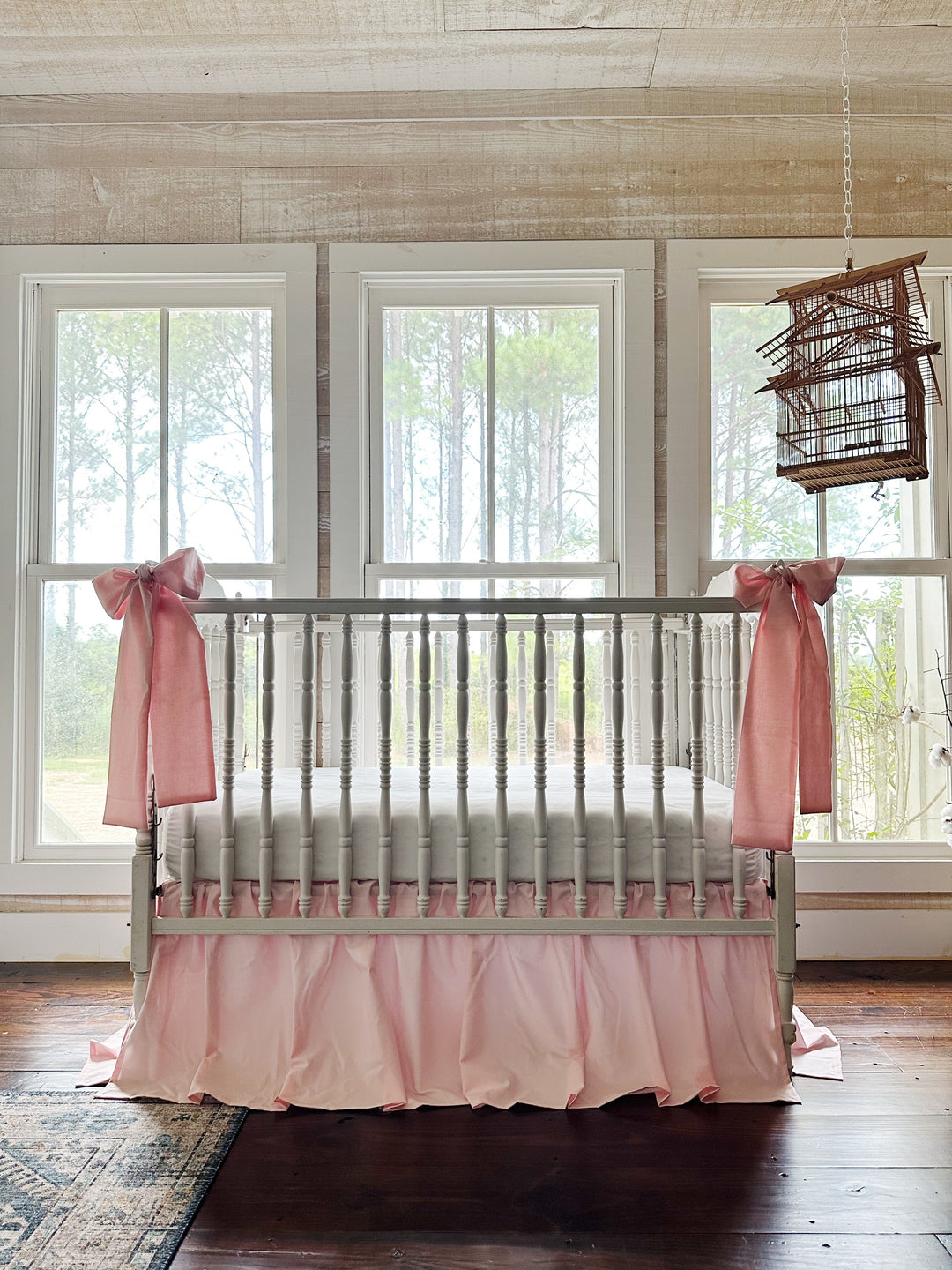 Sisters Pink crib skirt and bow set styled in a farmhouse nursery with pine views and a hanging birdcage