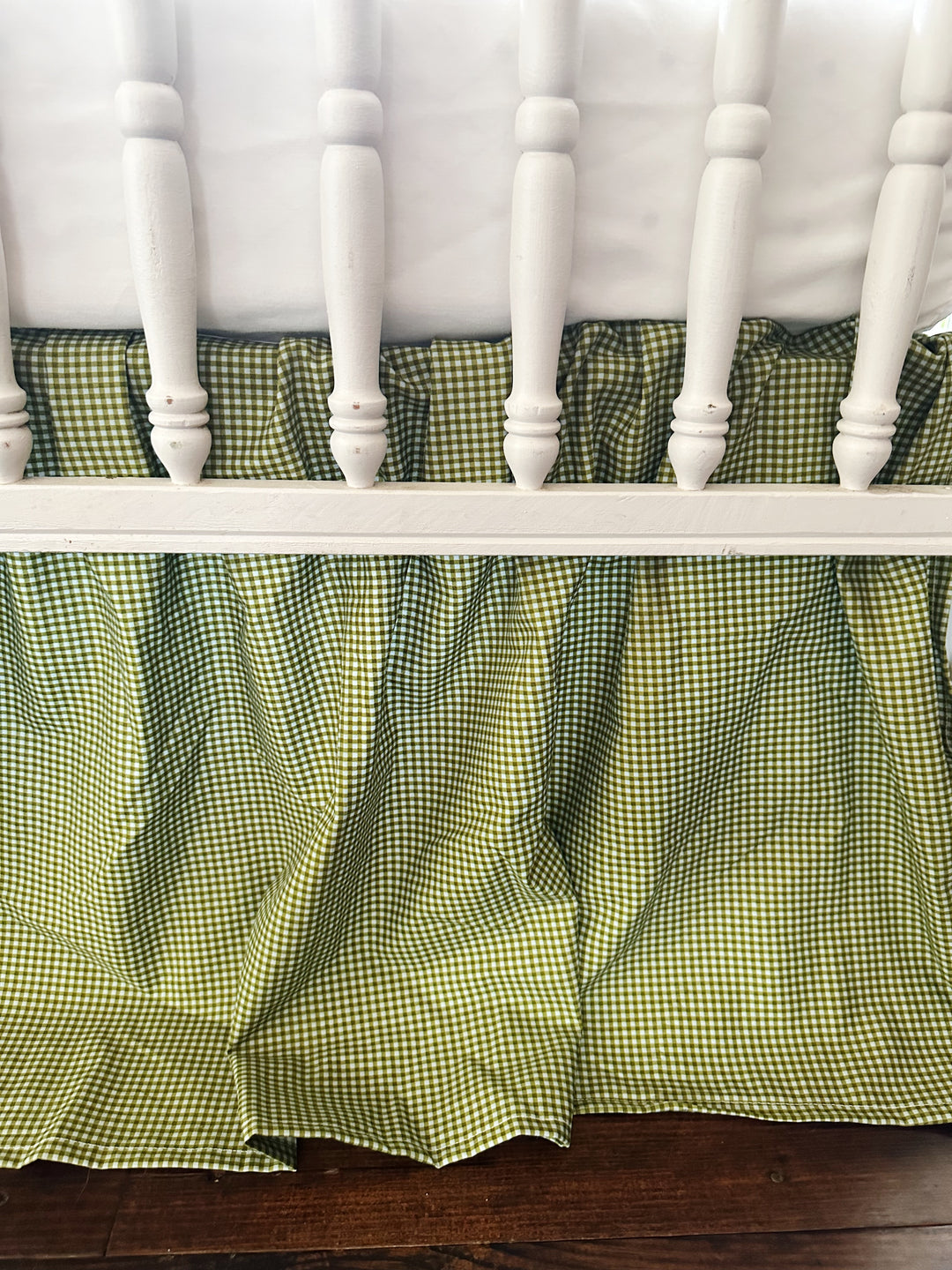 Gathered pleats of the pine green gingham farmhouse crib skirt behind white crib spindles