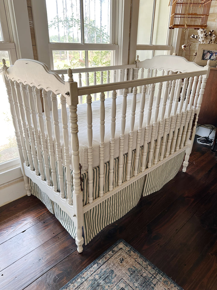 Pine green ticking tailored crib skirt on a white spindle crib, pleated panels falling to the floor