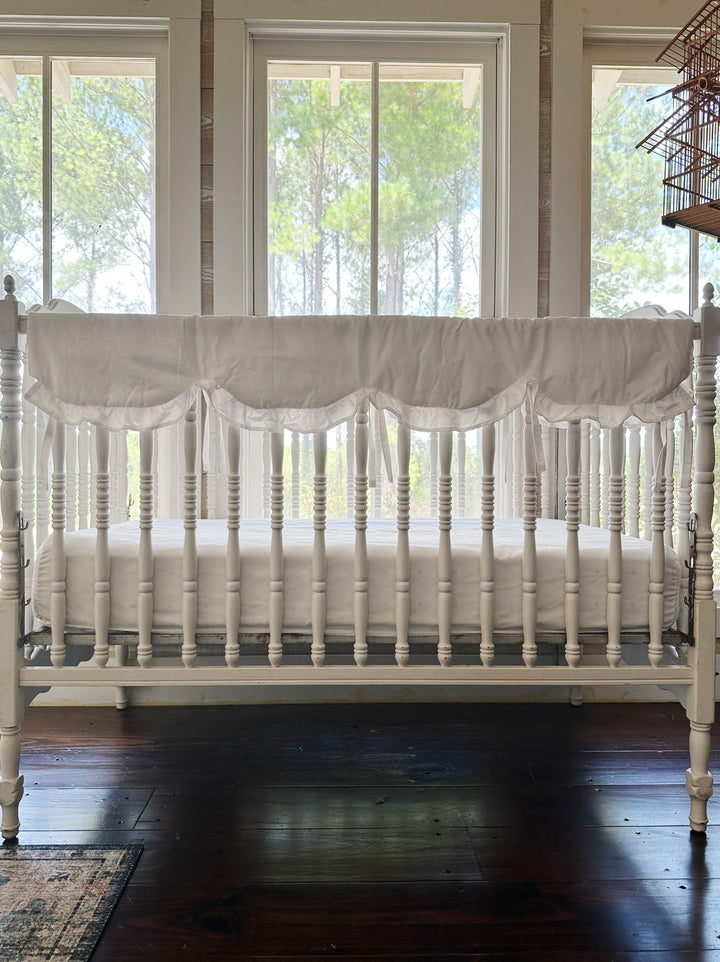White ruffled scalloped crib rail cover tied across the long top rail of a white spindle crib