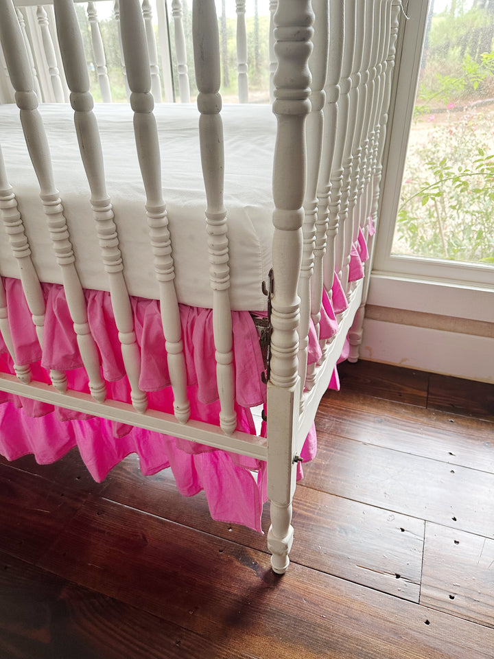 Corner of a white spindle crib where the pink ruffle tiers wrap from the side around to the end