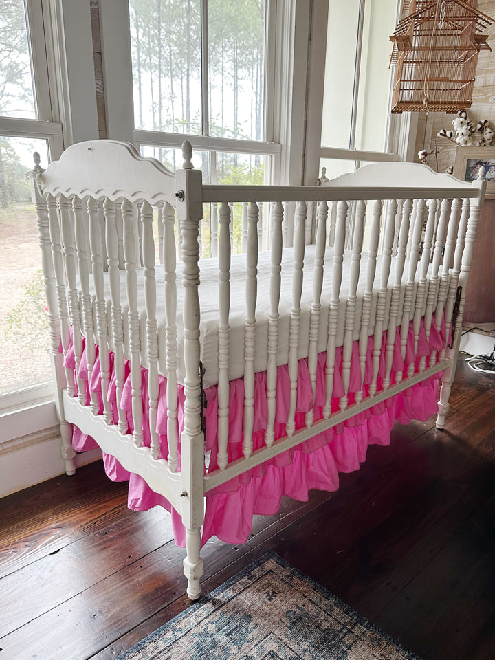 Three-quarter view of the tiered pink crib skirt on a white spindle crib by the windows, ruffles running along the side and foot