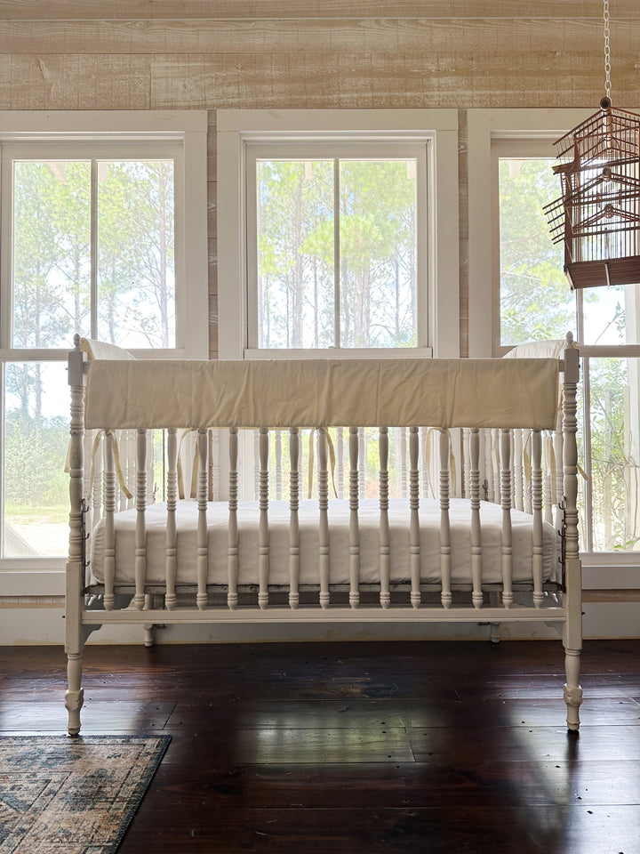 Nursery view of the ivory rail cover on a white crib beneath three windows framed by pines