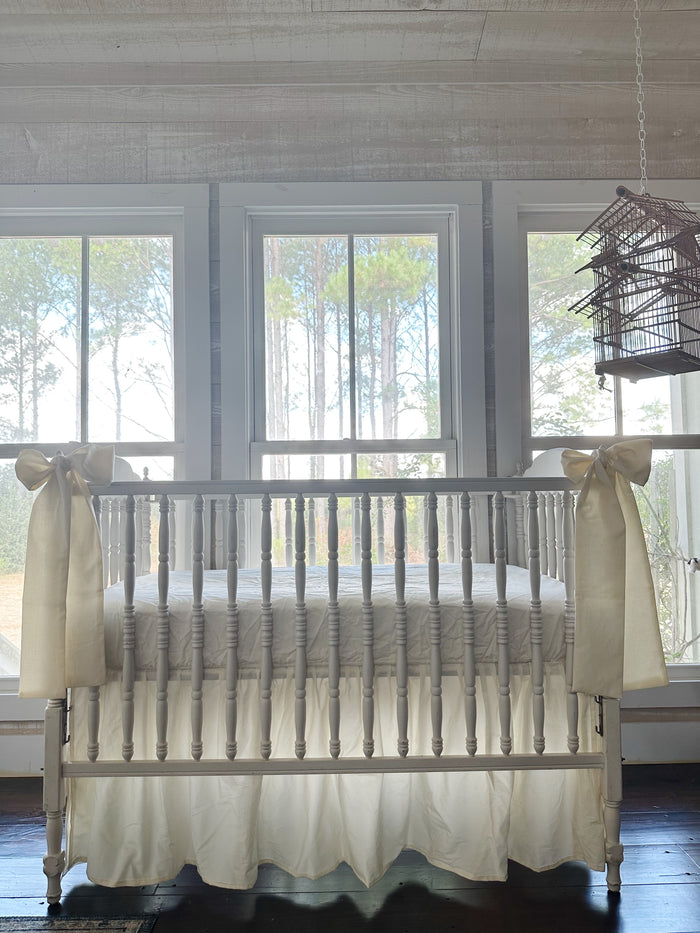Ivory farmhouse crib skirt and two ivory bows on a white spindle crib, straight-on in front of three tall windows
