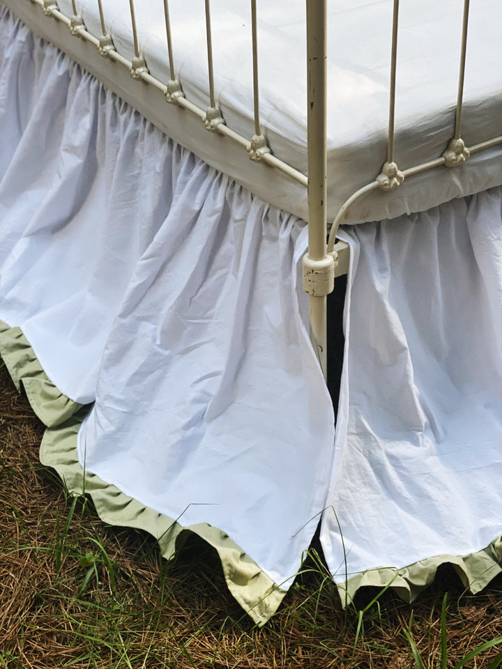 Corner detail of white gathered crib skirt with sage green ruffle hem and split corner around iron crib leg
