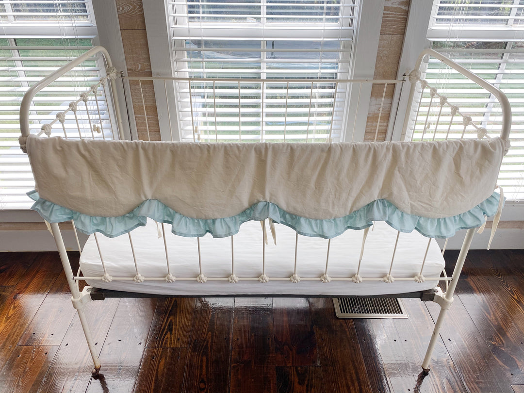 Ivory and mist scalloped crib rail cover shown across the long front rail of a vintage iron crib