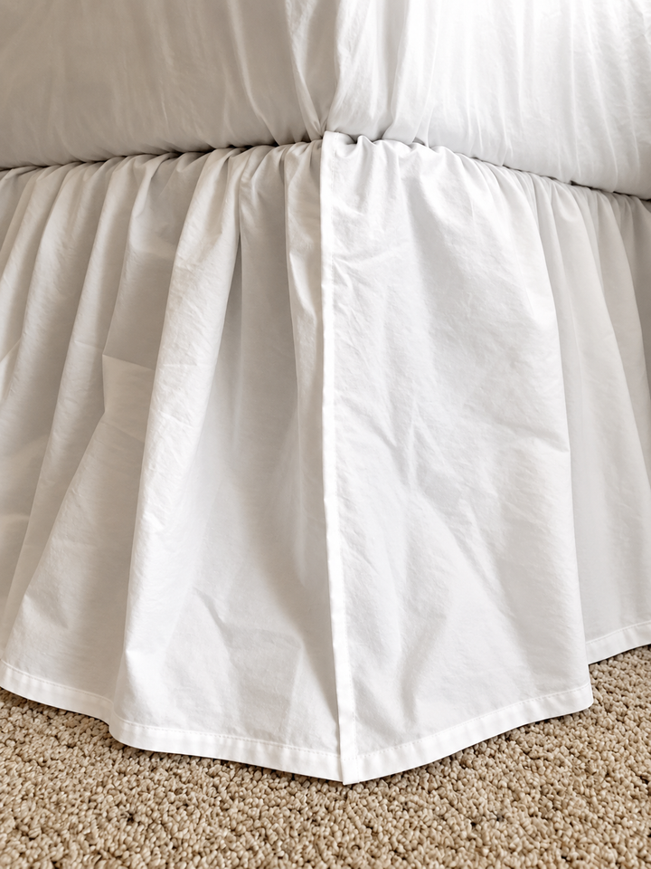 Close view of the white bed skirt corner over carpet, showing the gathered heading and the plain turned hem with no ruffle