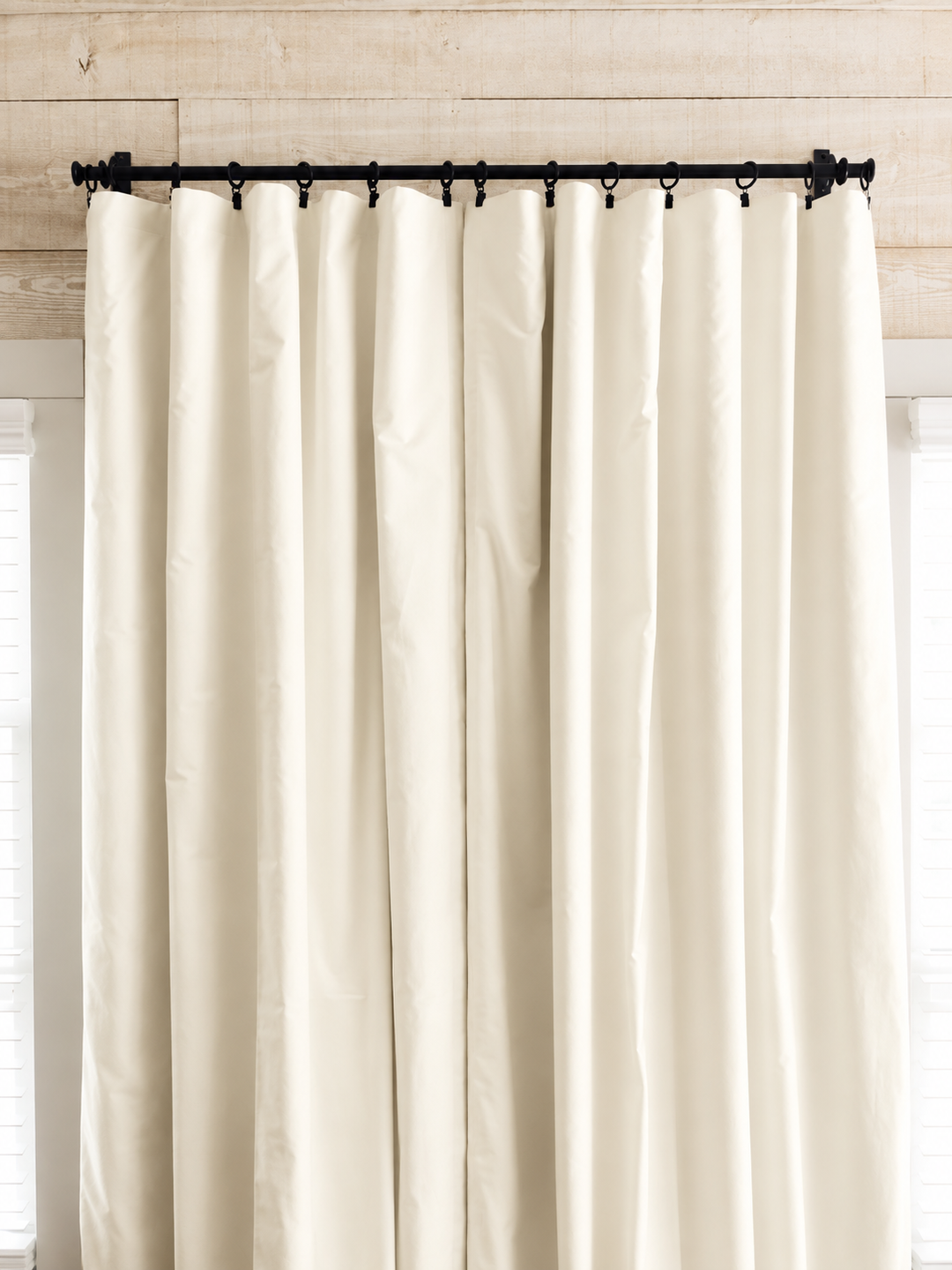 Ivory blackout curtain panels drawn closed on a black iron rod with clip rings, against a whitewashed plank wall