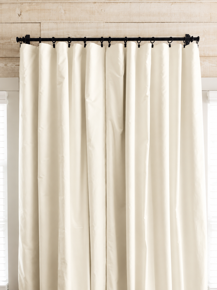 Ivory blackout curtain panels drawn closed on a black iron rod with clip rings, against a whitewashed plank wall