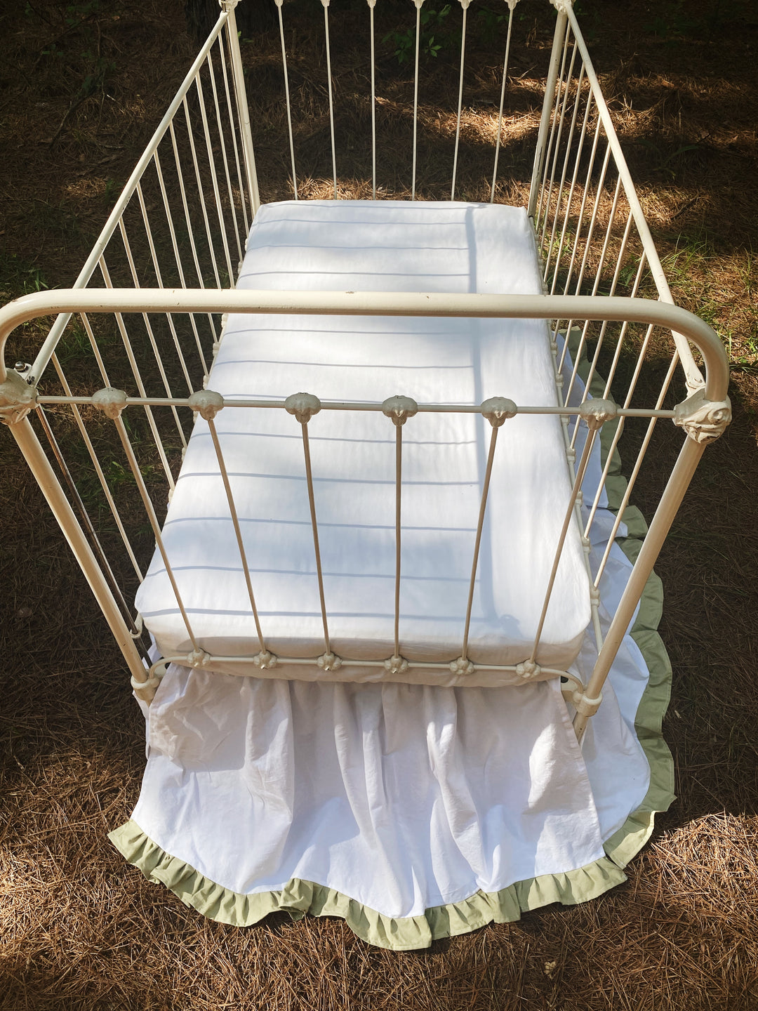 Bird's-eye view of white and sage ruffled crib skirt on iron crib outdoors, all-sided coverage visible