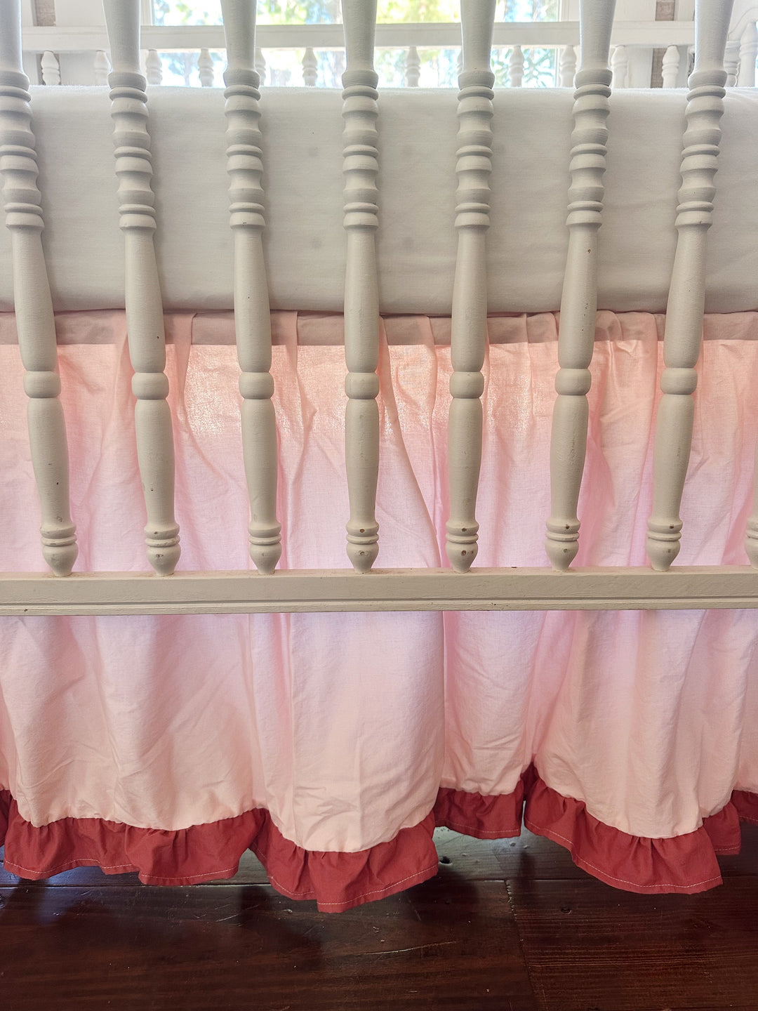Close-up through the crib spindles: gathered Sisters Pink cotton skirt ending in a blush ruffle just above the wood floor