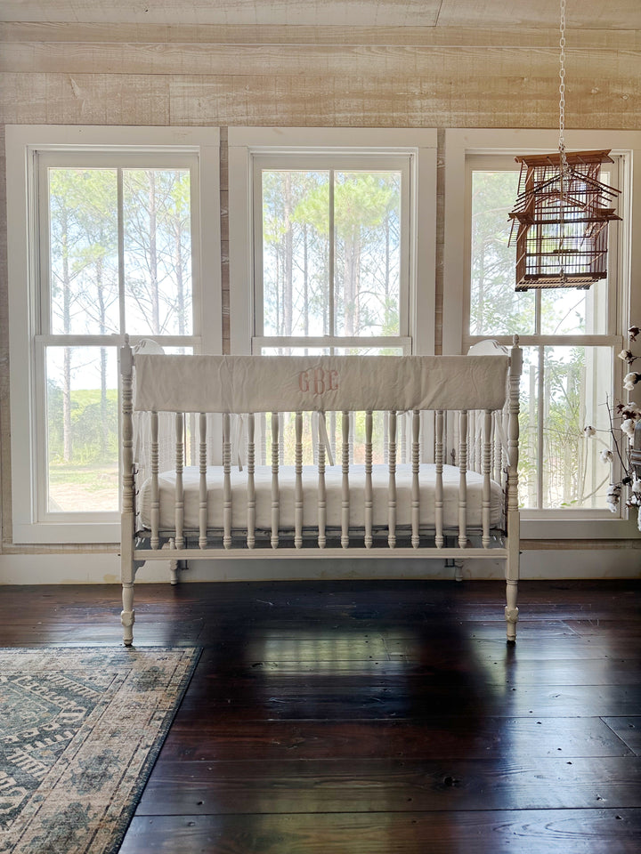 Wide nursery view of the monogrammed white rail cover on the crib above dark wood floors