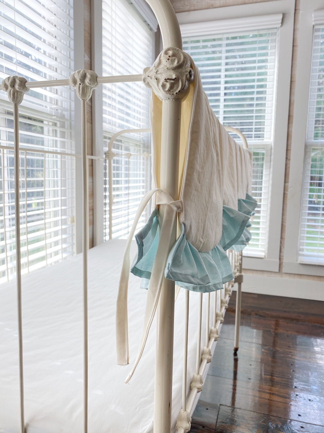 Ivory cotton tie knotted at the corner post of an iron crib beside the mist blue ruffle