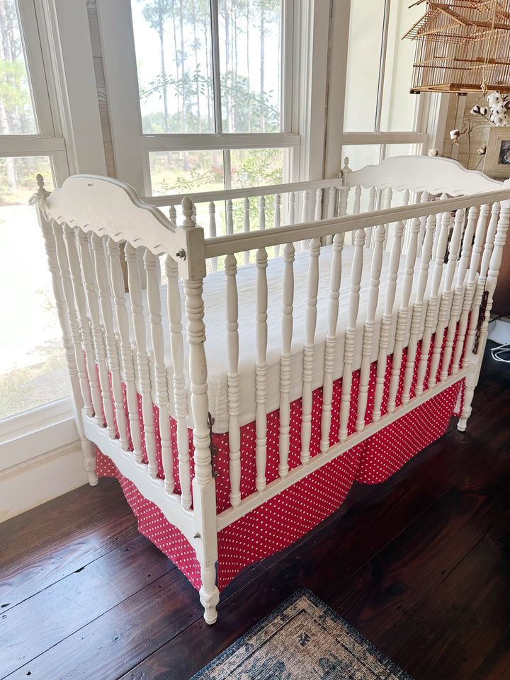 Three-quarter view of a white spindle crib wearing the cherry red and white polka dot tailored skirt