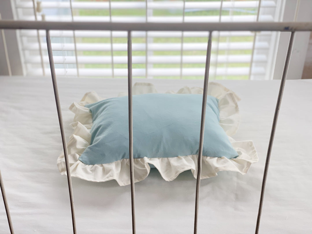 Mist crib pillow with ivory ruffle lying flat behind the bars of an iron crib
