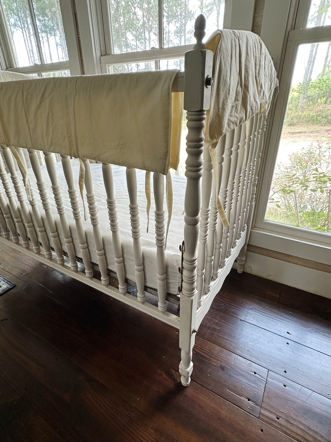 Ivory crib rail covers seen from the corner of the crib with windows and greenery behind