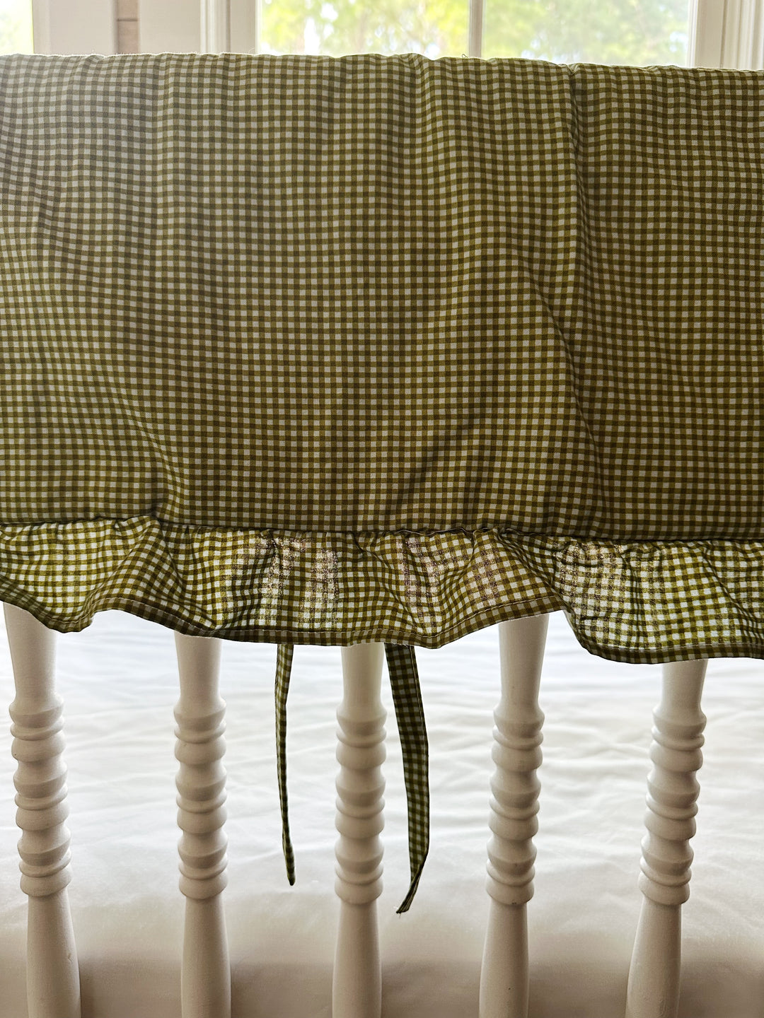 Ruffle edge and tiny fabric ties on the green gingham crib rail cover, backlit by a window