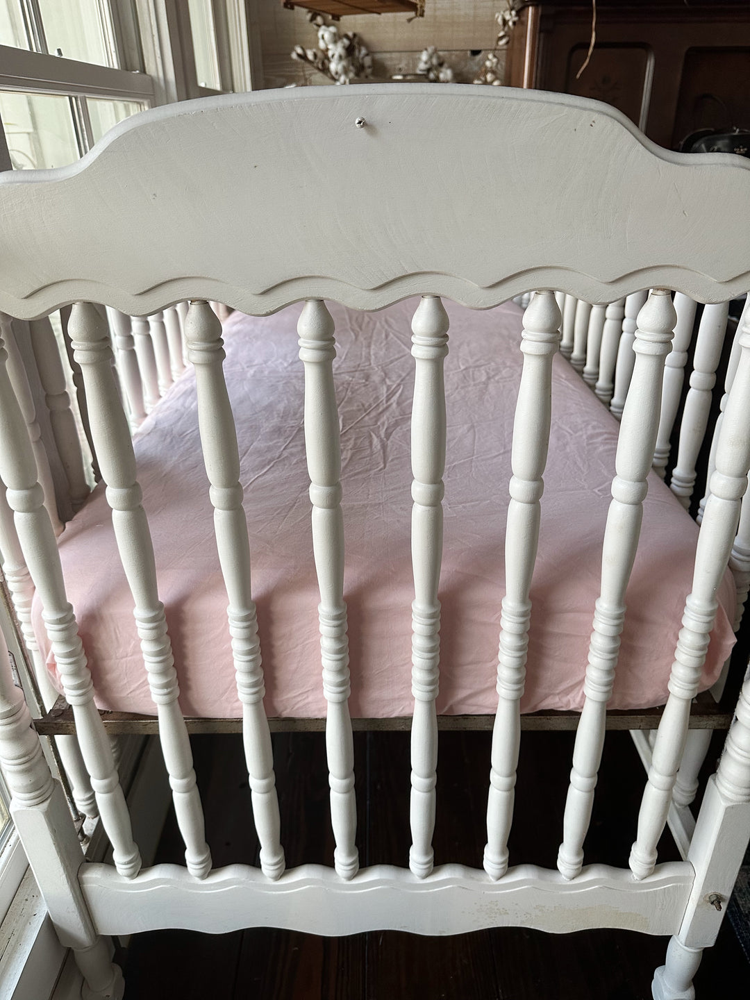 Foot end of the crib with the Sisters Pink fitted crib sheet wrapped smoothly over the mattress, seen through scalloped spindles