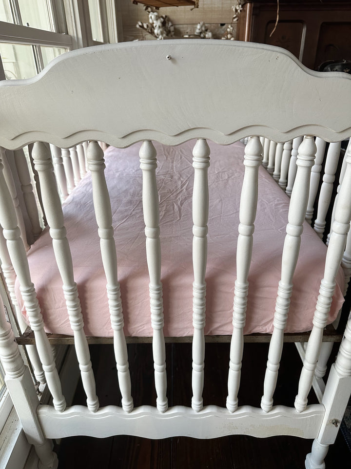 Foot end of the crib with the Sisters Pink fitted crib sheet wrapped smoothly over the mattress, seen through scalloped spindles