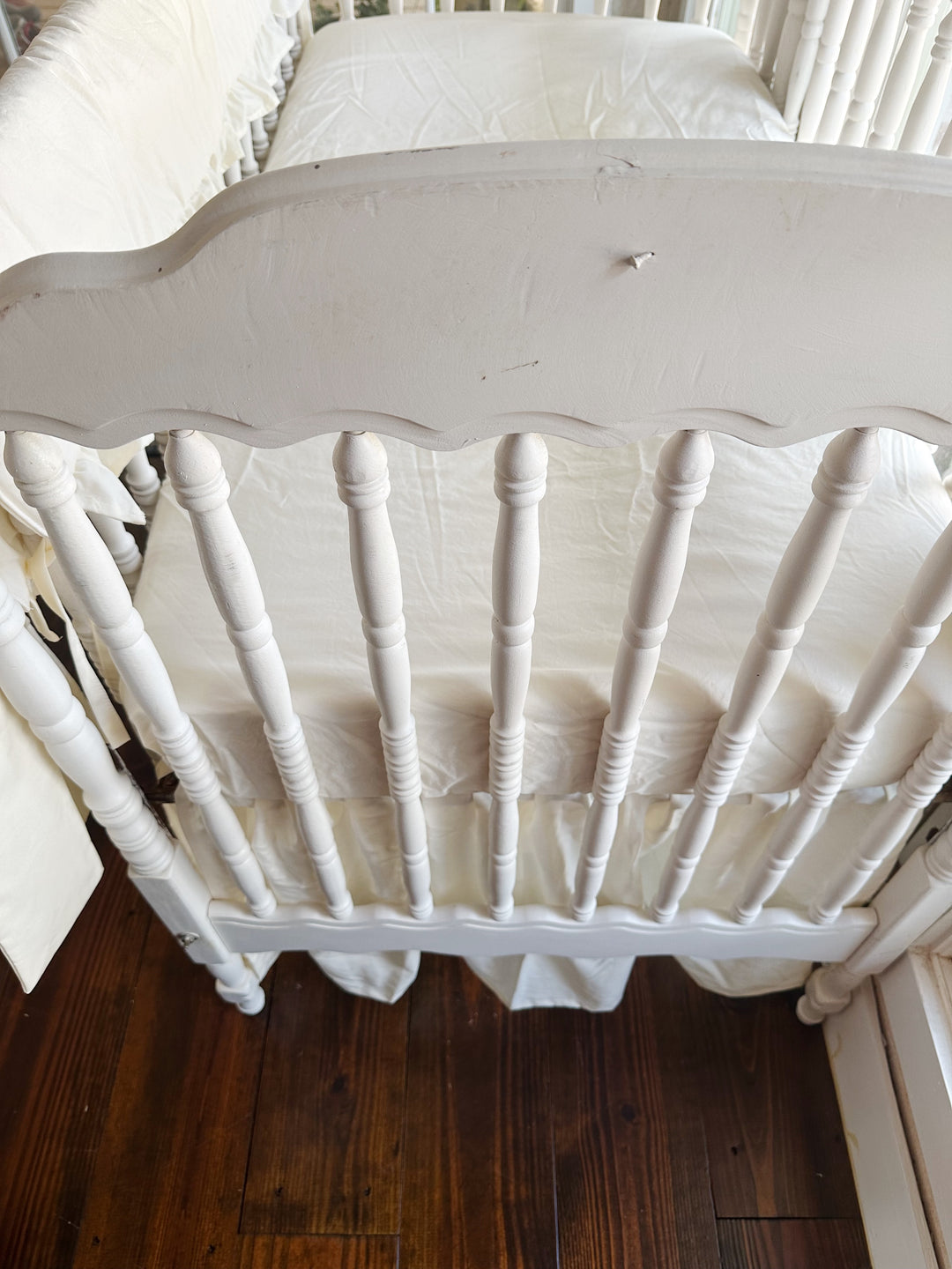 Looking down over the scalloped end of the crib at an ivory fitted mattress, gathered ivory skirt and the rail cover along the side