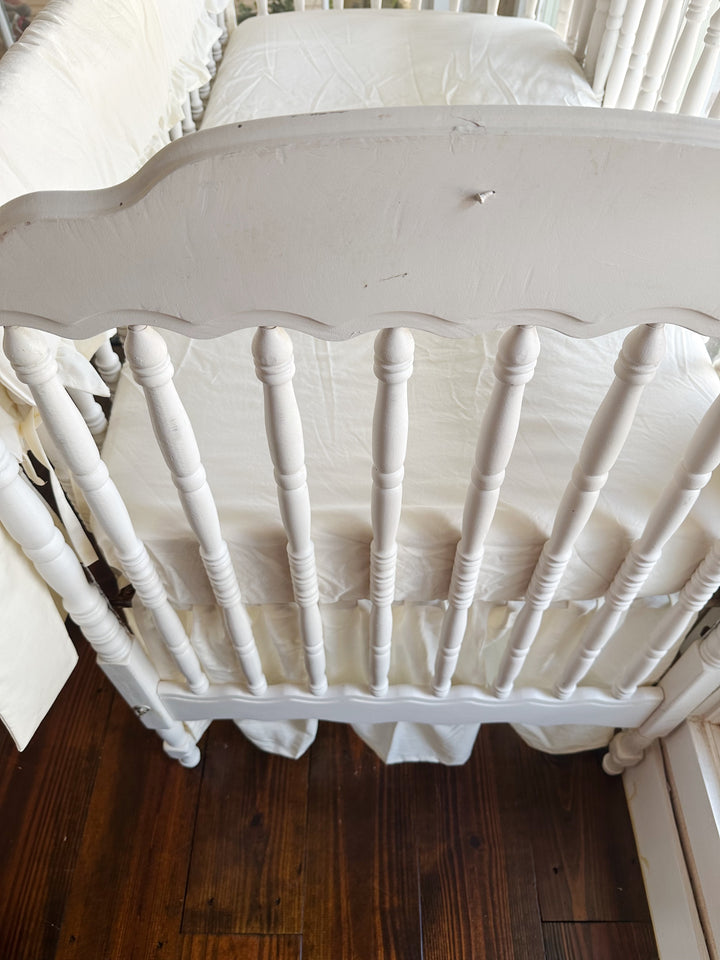 Looking down over the scalloped end of the crib at an ivory fitted mattress, gathered ivory skirt and the rail cover along the side