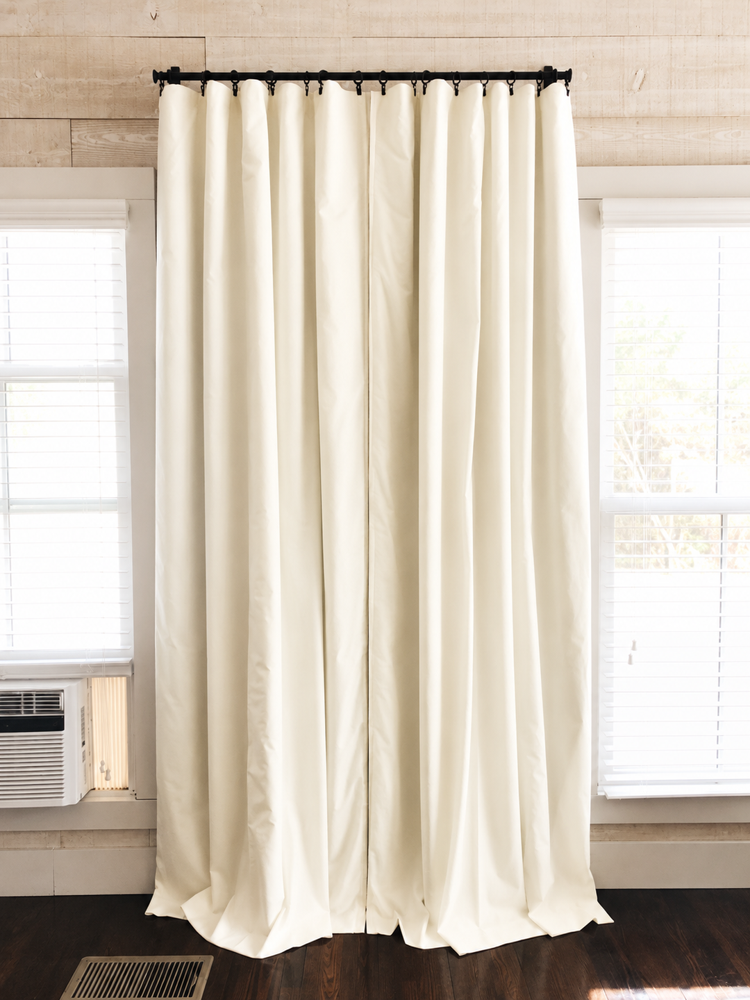 A pair of ivory blackout panels closed across a window, hanging rod to floor in a room with dark pine floors