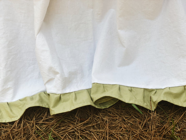 White and sage ruffled crib skirt three-quarter angle view in woodland setting, full gathered silhouette on iron crib