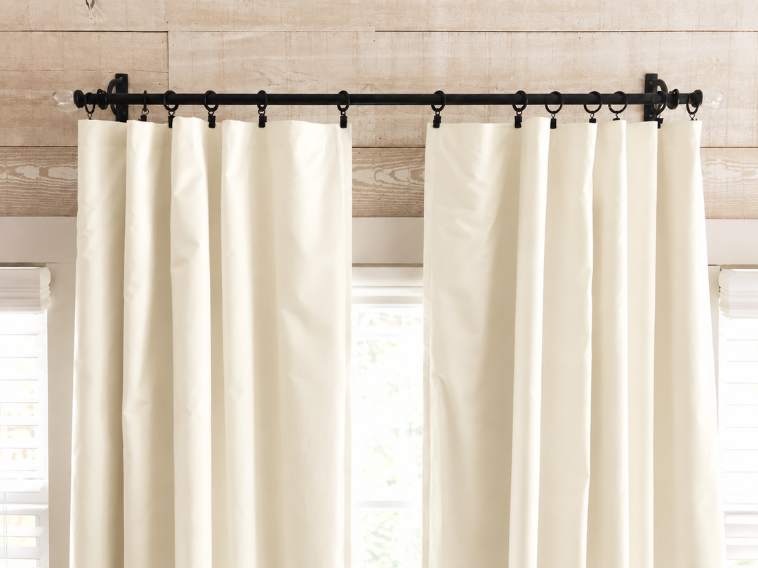 Ivory panels parted on a black rod with rings, daylight and greenery showing through the window between them