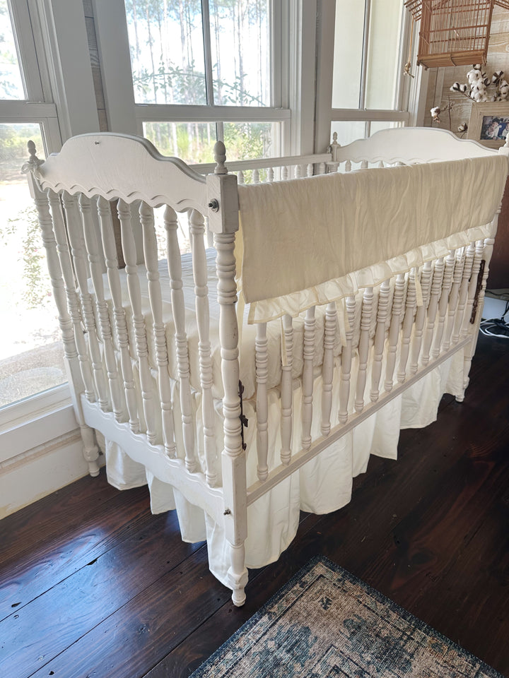 Three-quarter view of the ivory rail cover and skirt set on a white spindle crib beside the windows, skirt split around the corner leg
