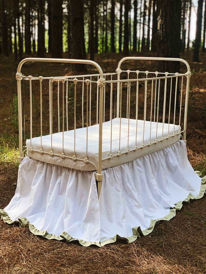 Front full view of white gathered crib skirt with sage ruffle trim on iron crib in dappled forest light, heirloom nursery