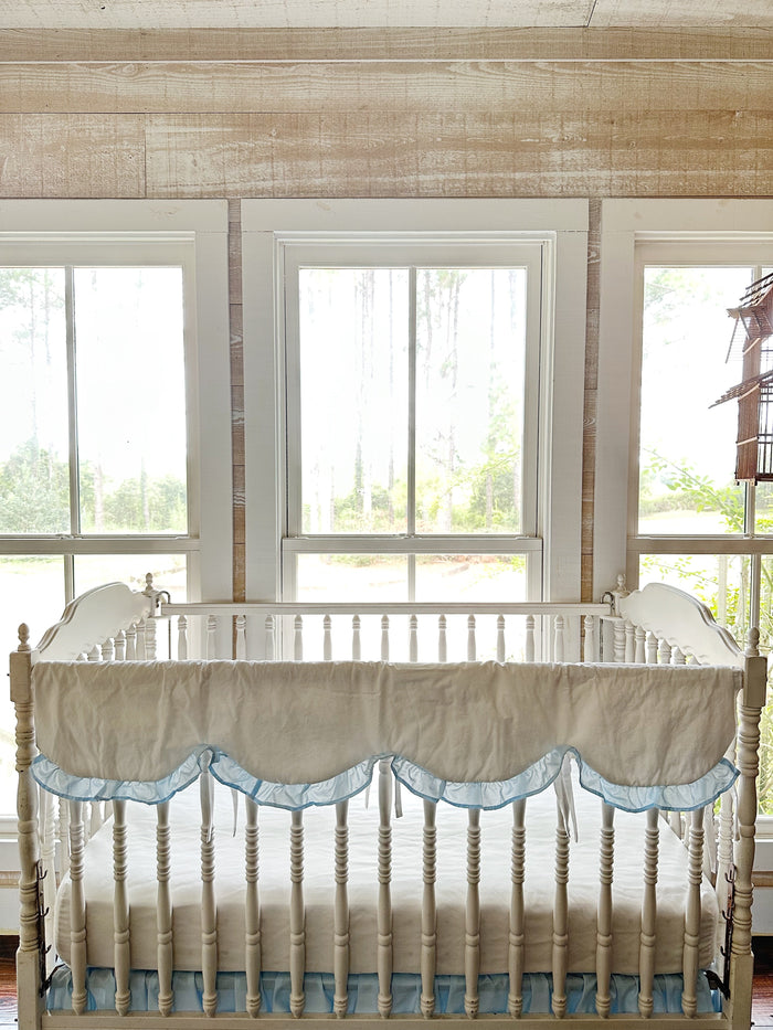 Scalloped rail cover on a white crib framed by tall windows and pine woods beyond